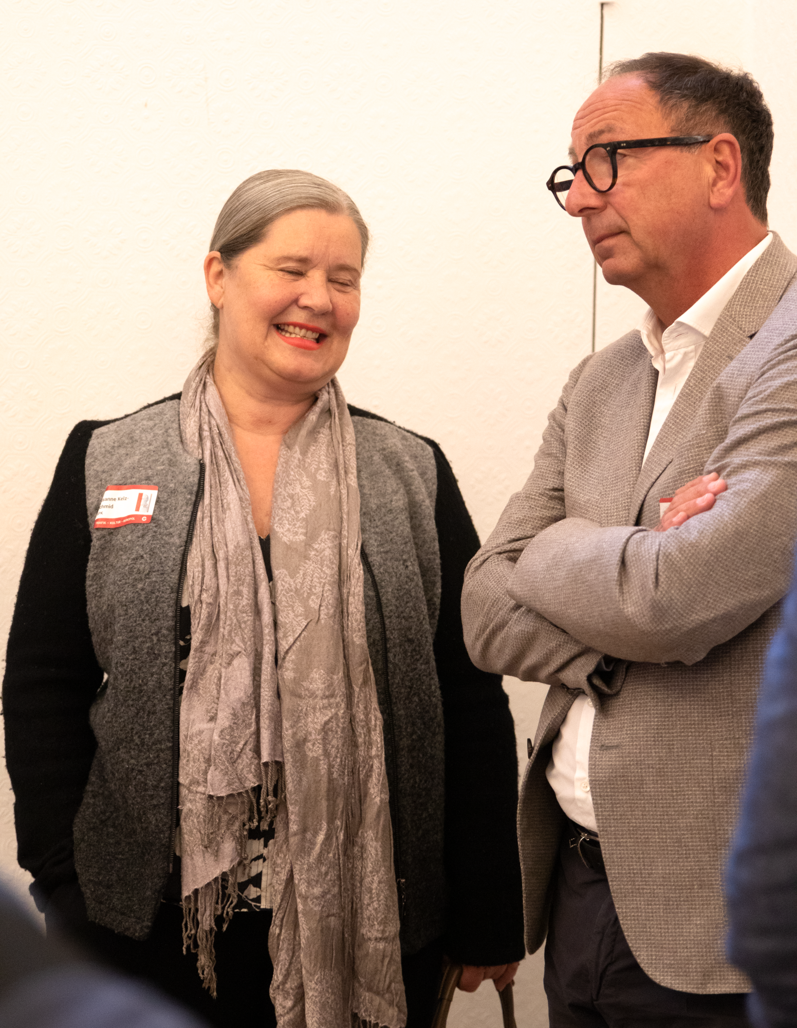 Eine Frau mit einem beigen Schal und einer grau schwazen Jacke steht neben einem Herren mit einem weißem Hemd und einem braunen, grauen Sakko 