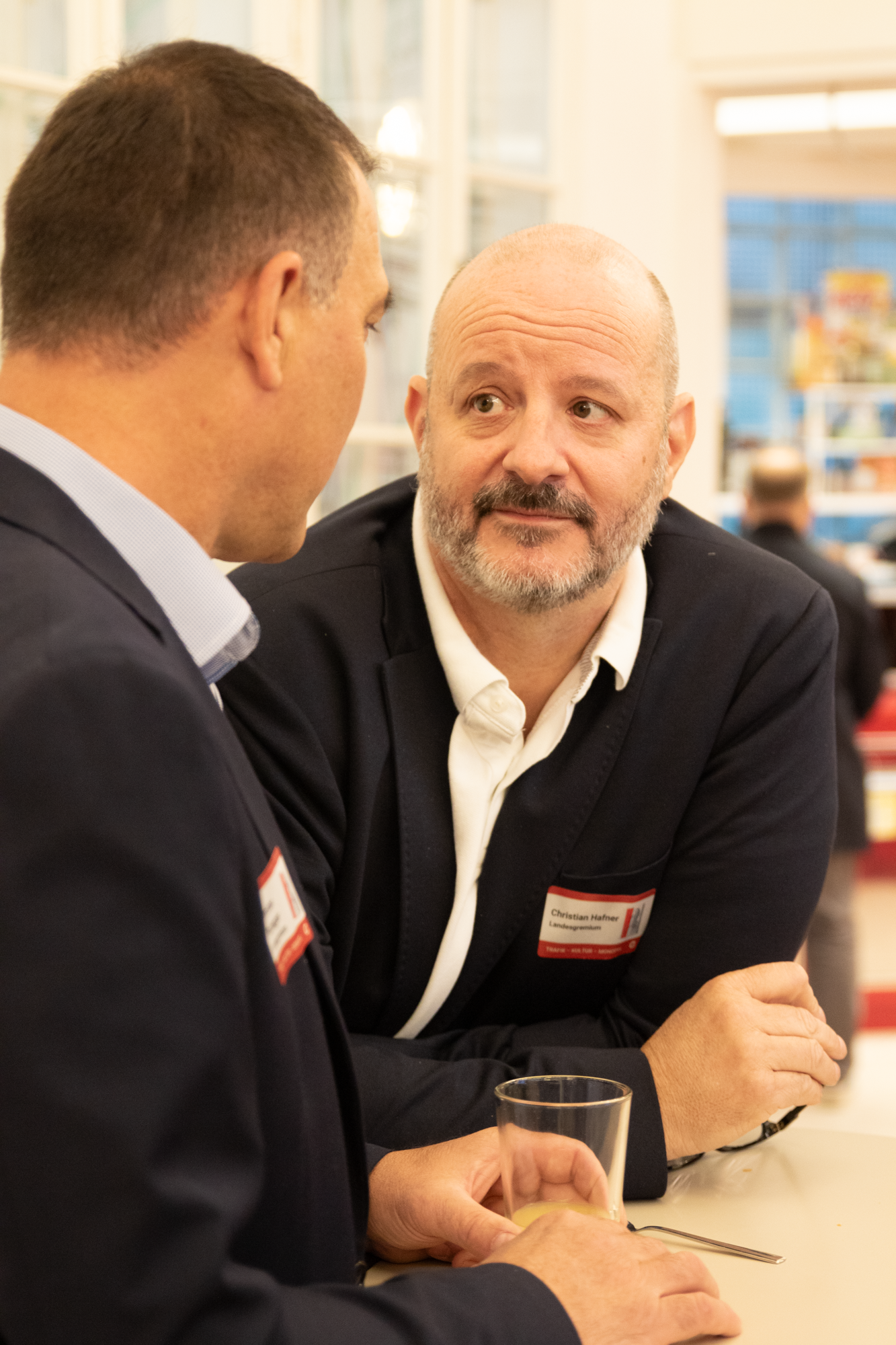 Ein Mann mit einem weißen Hemd und einem schwarzen Sakko nehnt sich an einem Tisch wo er sich mit einem Mann mit blauen Hemd und schwarzen Sakko unterhält