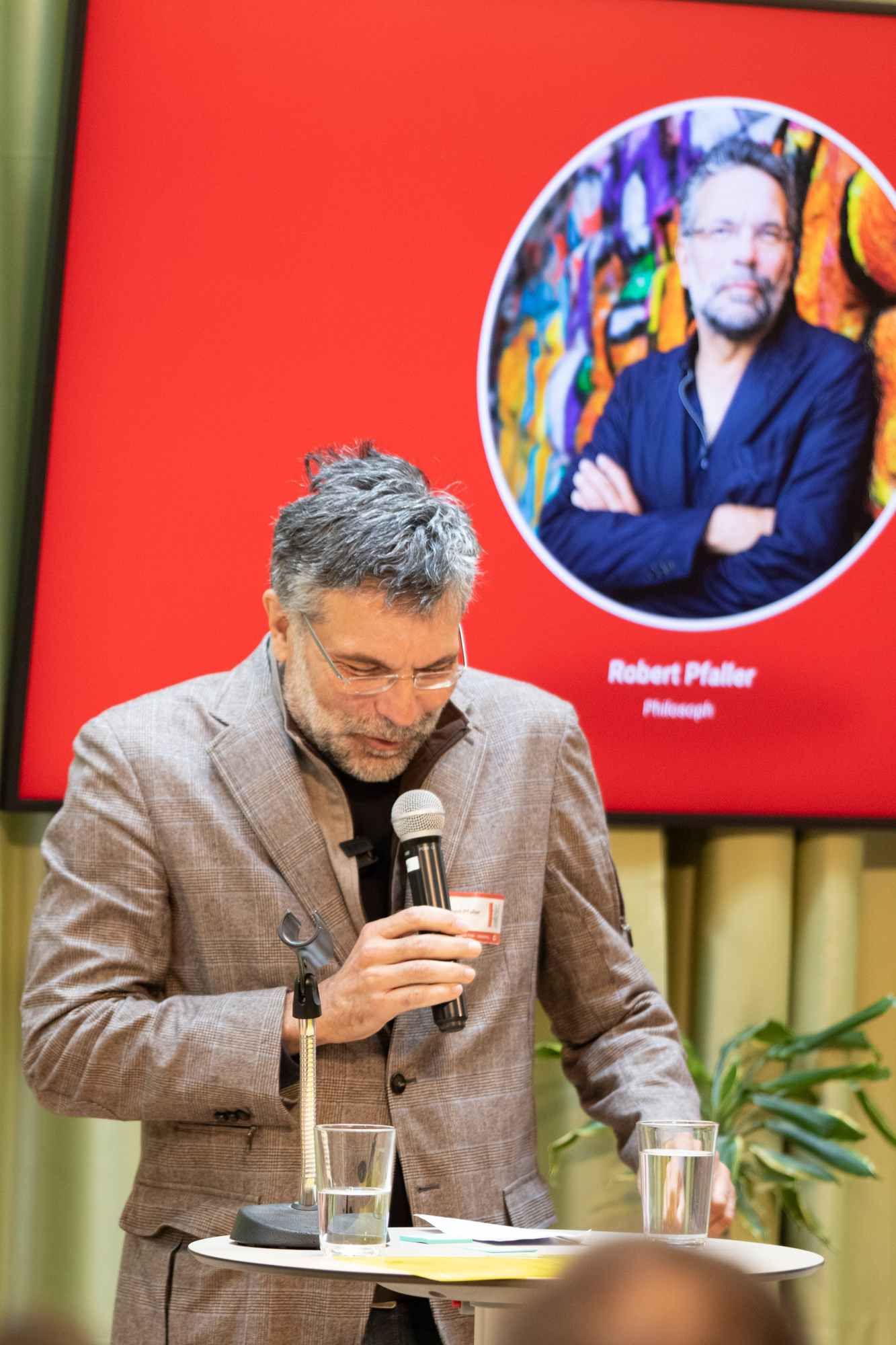 Ein älterer Herr steht auf der Bühne und präsentiert in einem braun, grauen Sakko das Trafikbuch