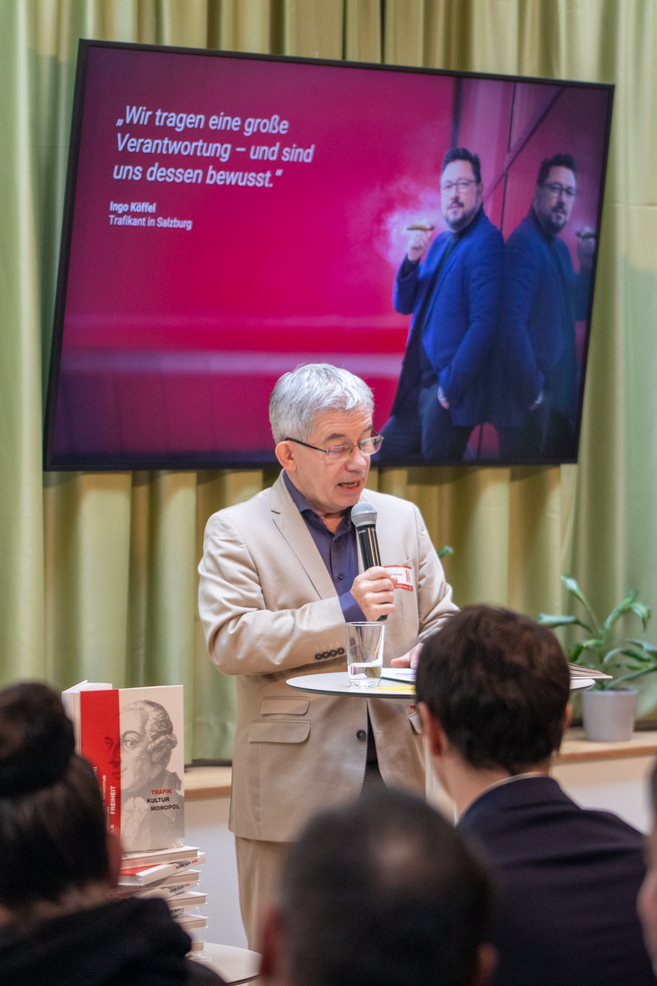 Monopolstellenleiter trägt einen beigen Anzug mit violetten Hemd auf der Bühne und präsentiert das Trafikbuch