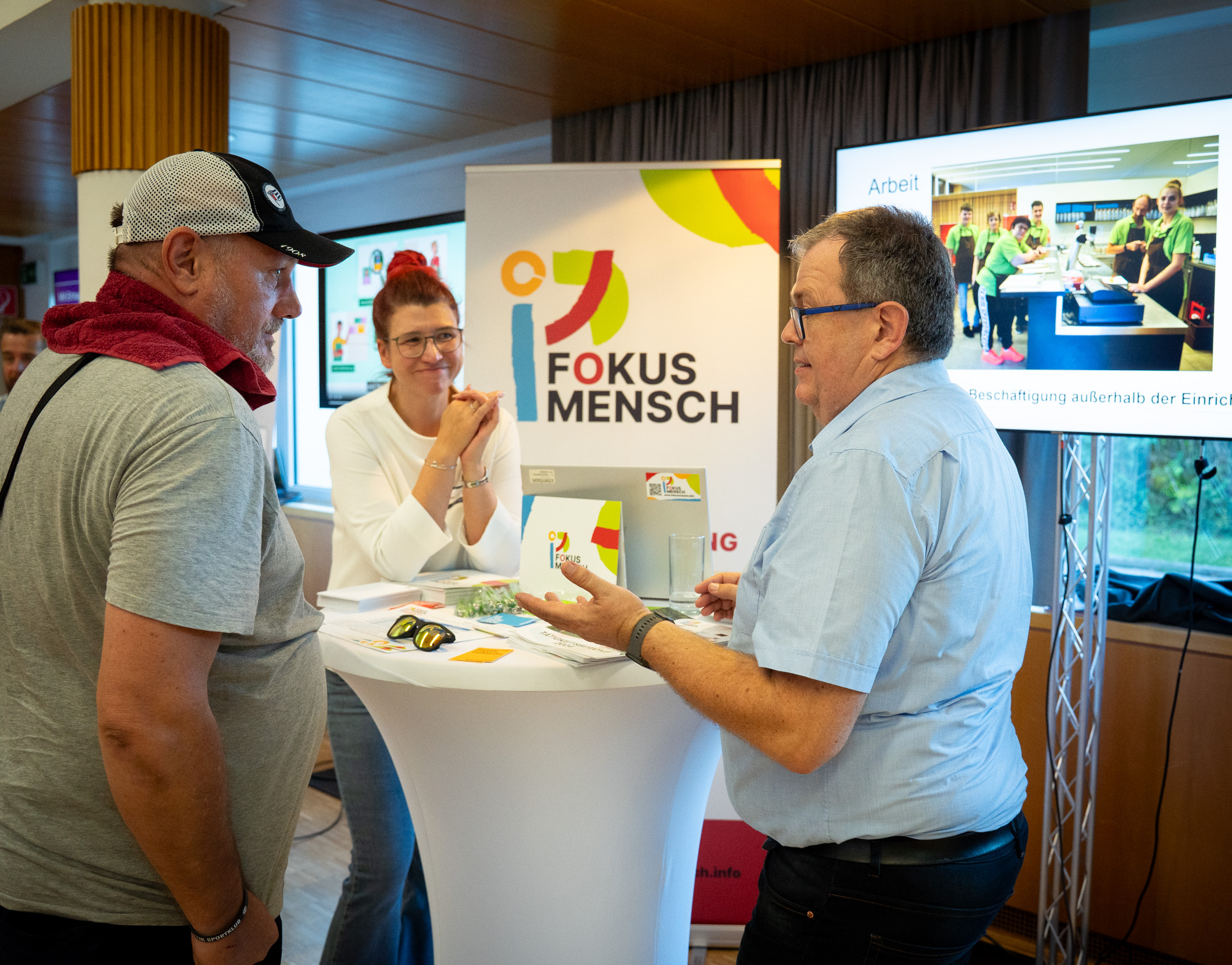 Drei Personen befinden sich auf diesem Bild, darunter sind zwei Männer und eine Frau. Ein Mann und eine Frau informieren einen Interessenten über ihre Organisation Fokus Mensch