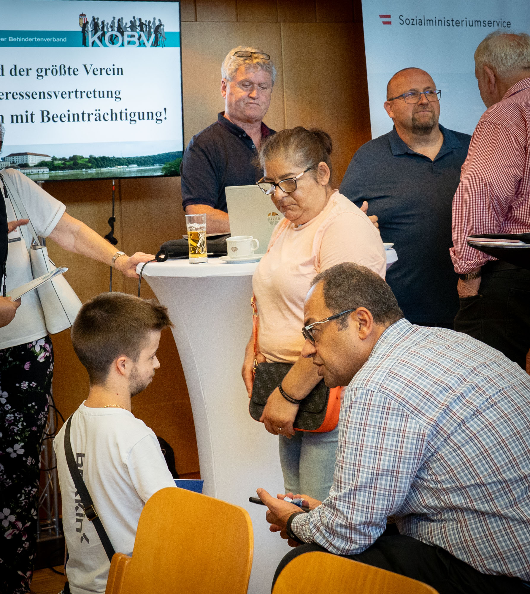 Im Hintergrund befinden sich Infostände von Organisation. Im Vordergrund unterhalten sich drei Personen. Eine Frau und zwei Männer. 