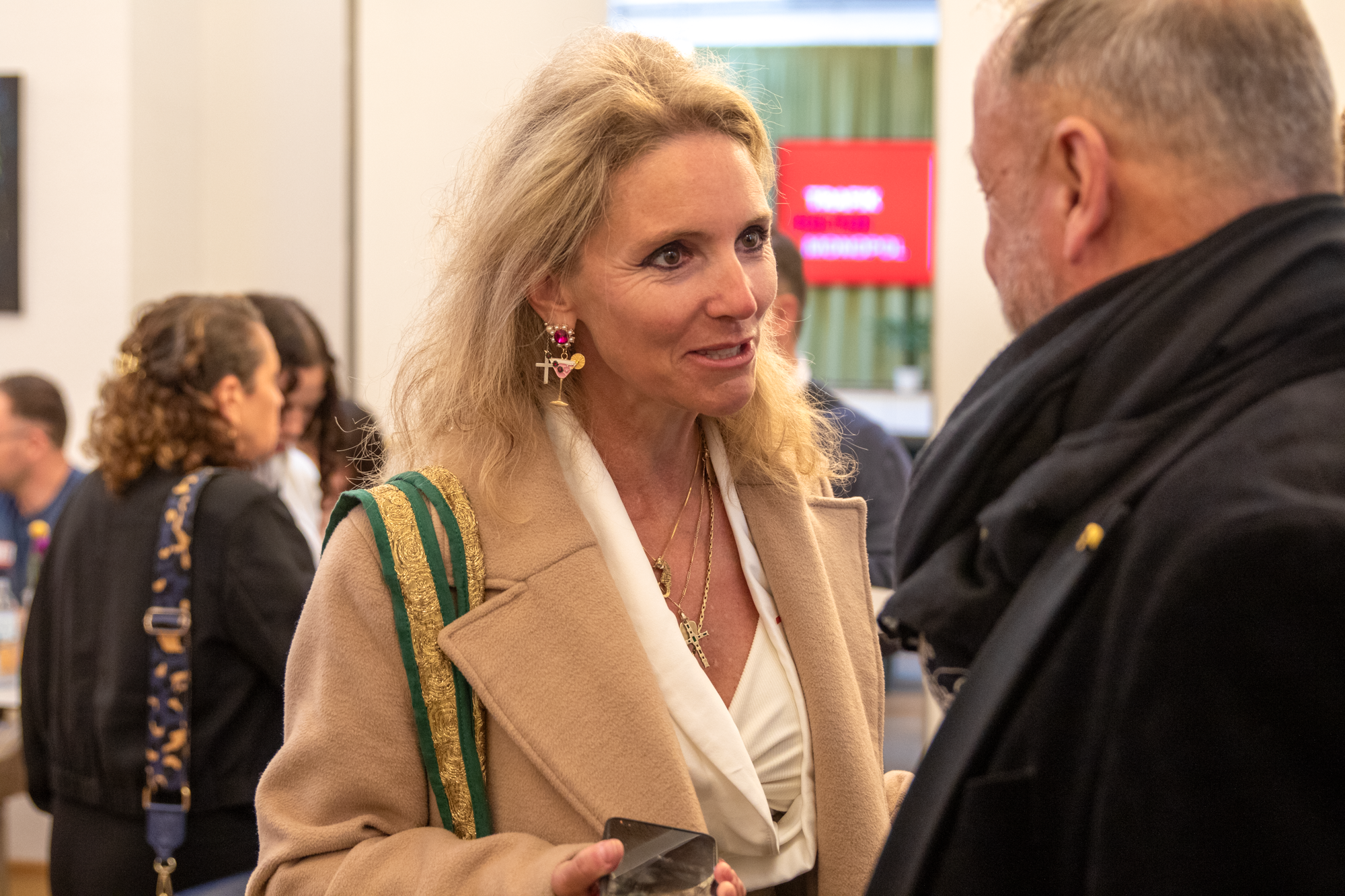 Eine blonde Frau mit einem beigen Mantel und einem weißen Oberteil unterhaltet isch mit einem Herren in einer schwarzen Jacke