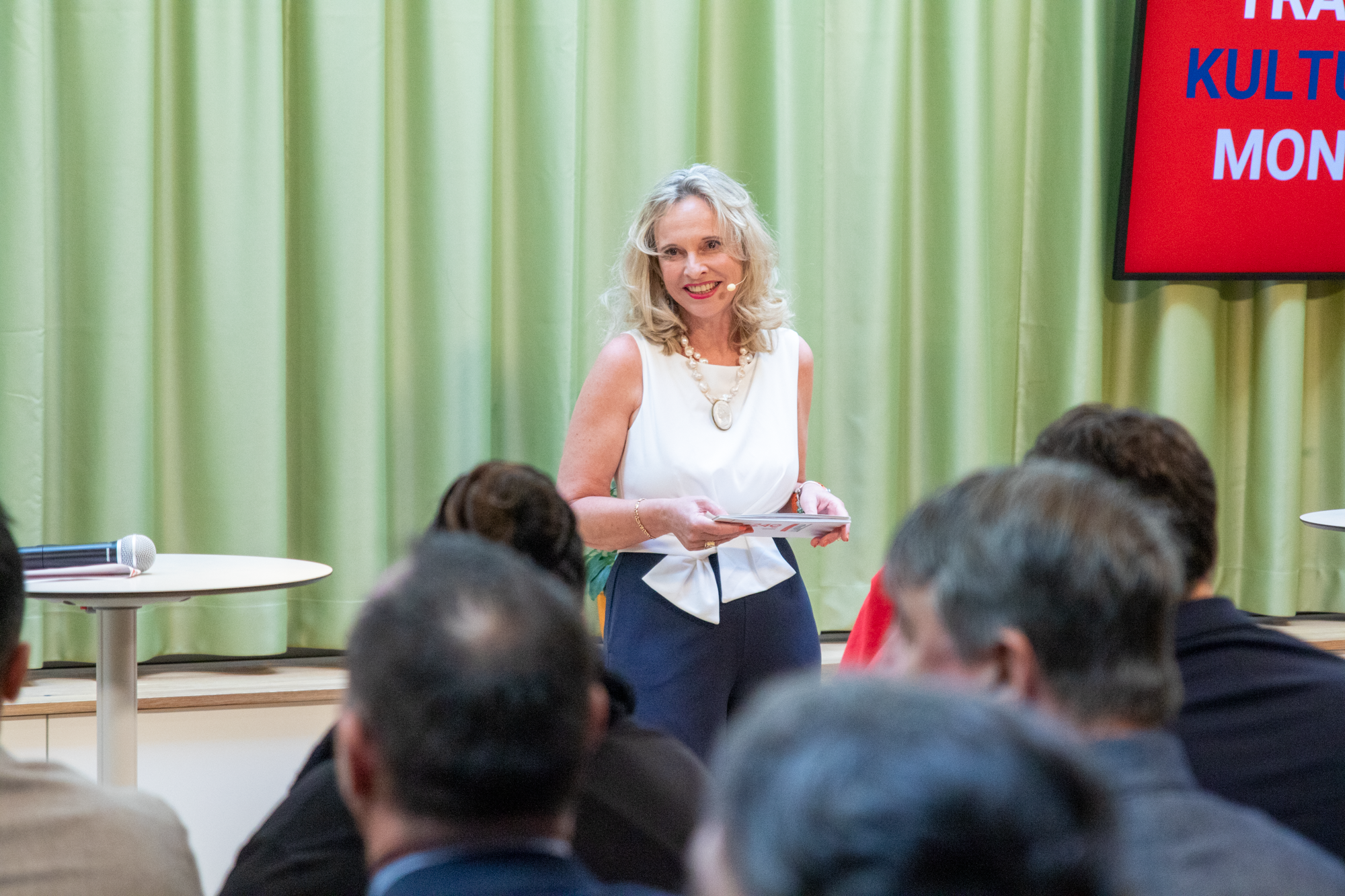 Monopolstellenleiterin moderiert in einem weißen Oberteil mit blauer Anzugshose