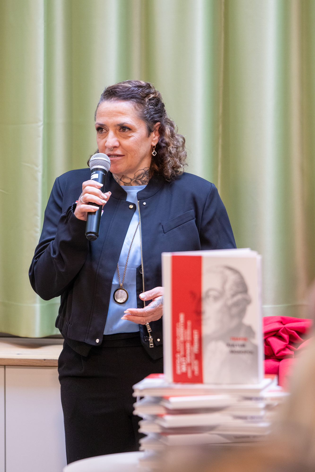 Eine Trafikantin mit einem hellblauen Oberteil, einer dunkelblauen Jacke und schwarzen Hose präsentiert Ihre Geschichte, welche im Buch beschrieben wird