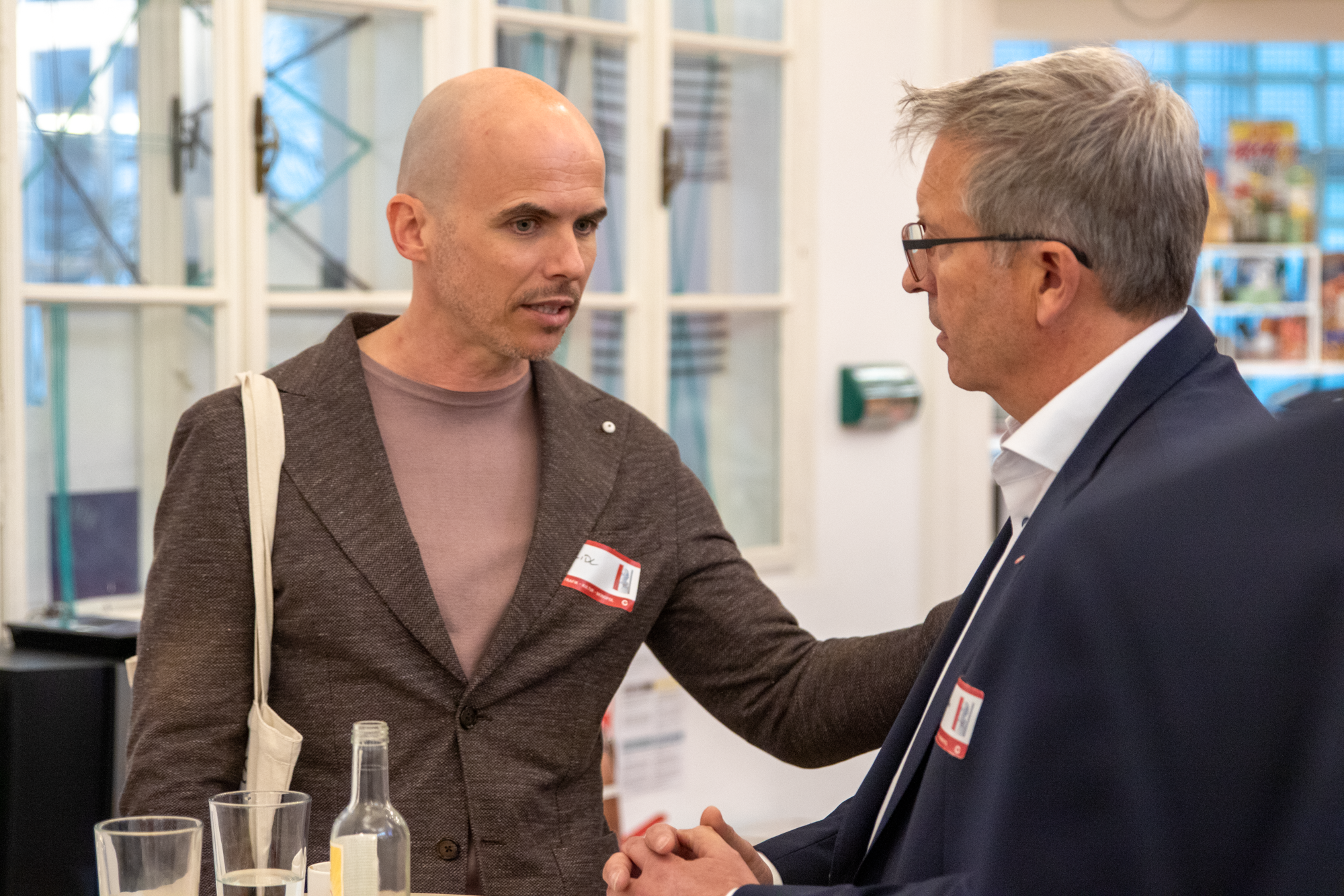Ein Mann mit braunem Sakko und einem dunkelrosa Pullover unterhaltet sich mit einen Monopolstellenleiter