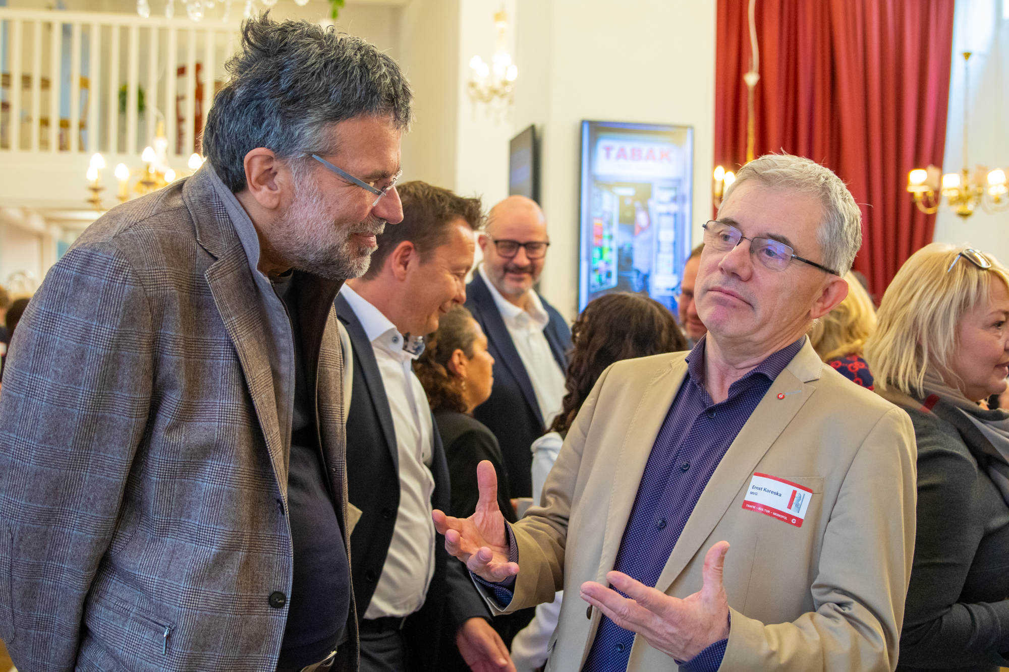 Monopolstellenleiter in beigen Sakko und violetten Hemd redet mit einem Besucher der Buchaustellung mit grauen Sakko und schwarzen Pullover