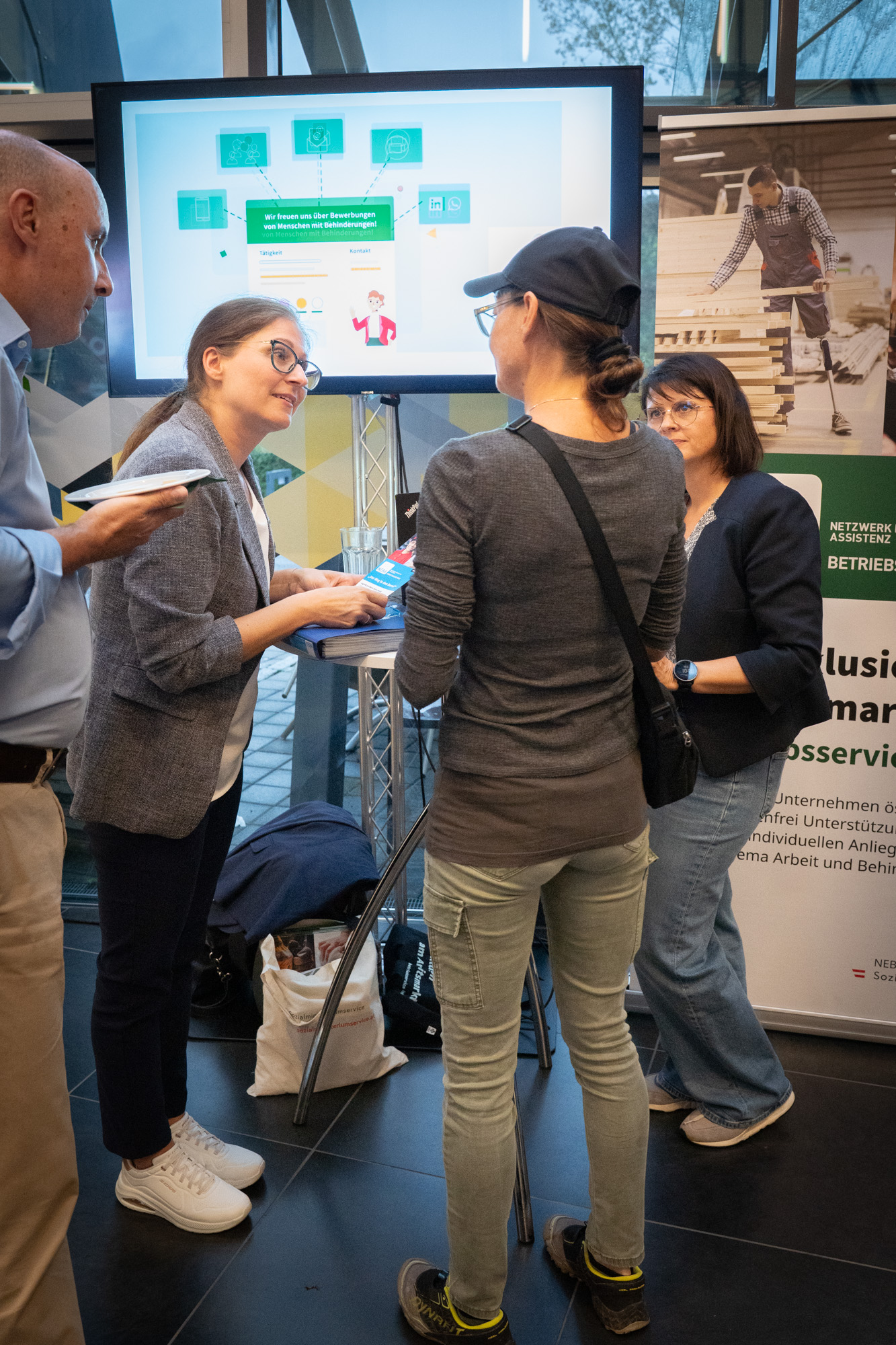 Vier Personen befinden sich auf dem Bild, drei Frauen und ein Mann. Eine Dame informiert sich bei einem Stand mit zwei Damen über deren Organisation. Ein Mann lauscht dem Gespräch.