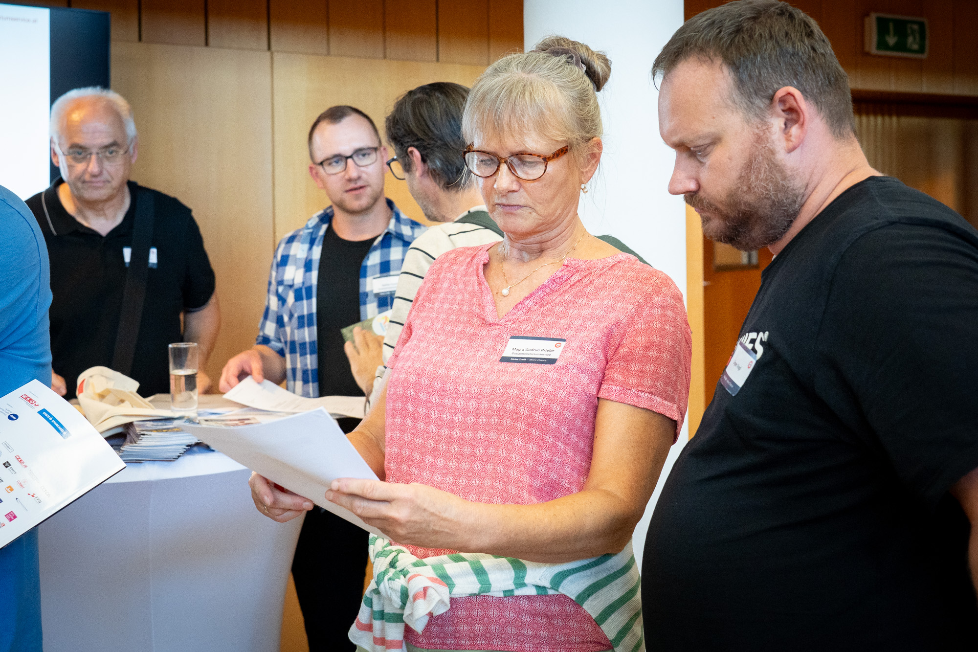 Im Hintergrund befindet sich ein Infostand wo sich mehrere Personen unterhalten. Im Vordergrund befindet sich eine Interessentin und ein Interessent lesen sich gemeinsam ein Infoblatt durch