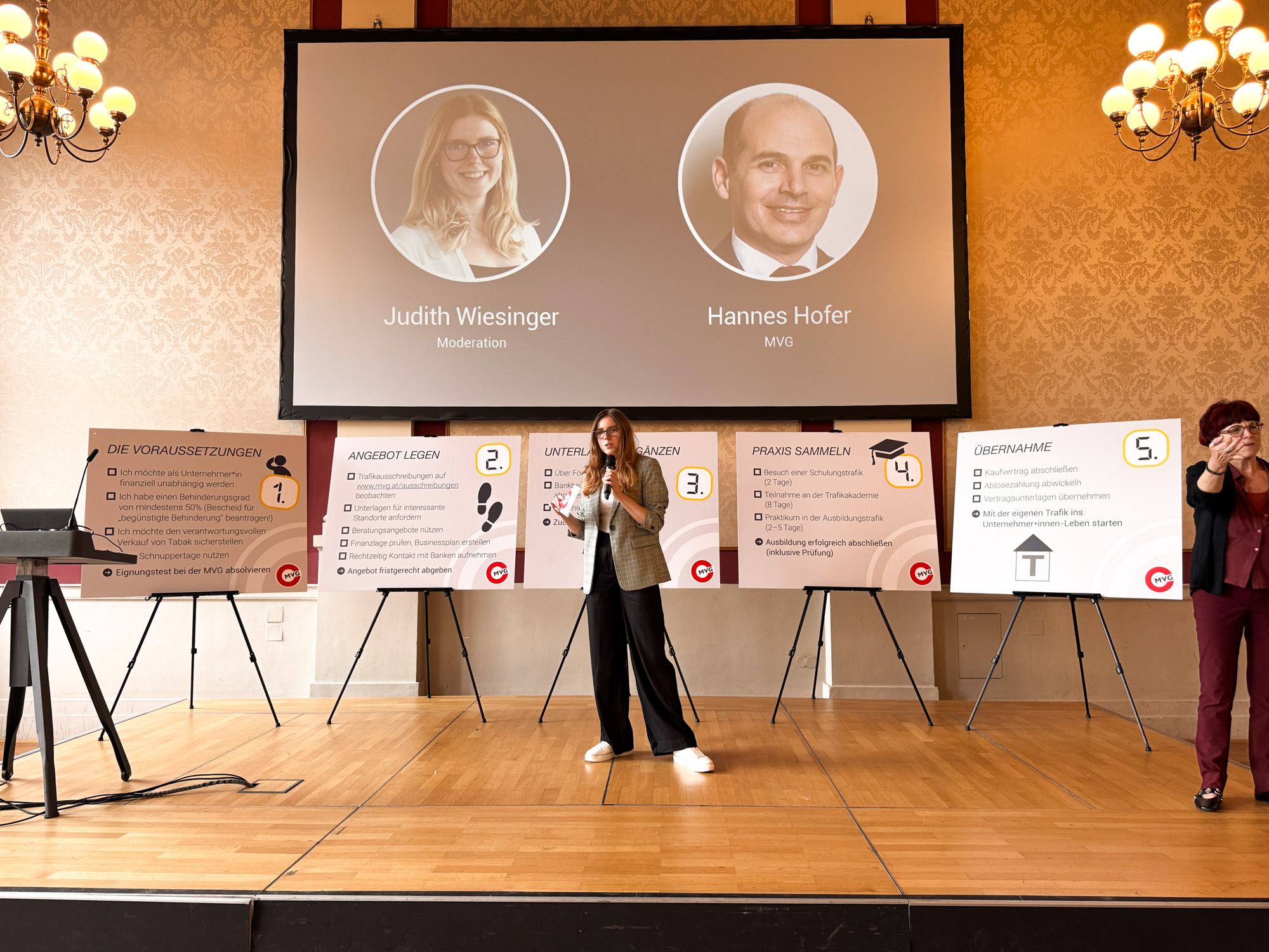 Die Moderatorin der Veranstaltung steht mit einer Gebärdensprachdolmetscherin auf der Bühne