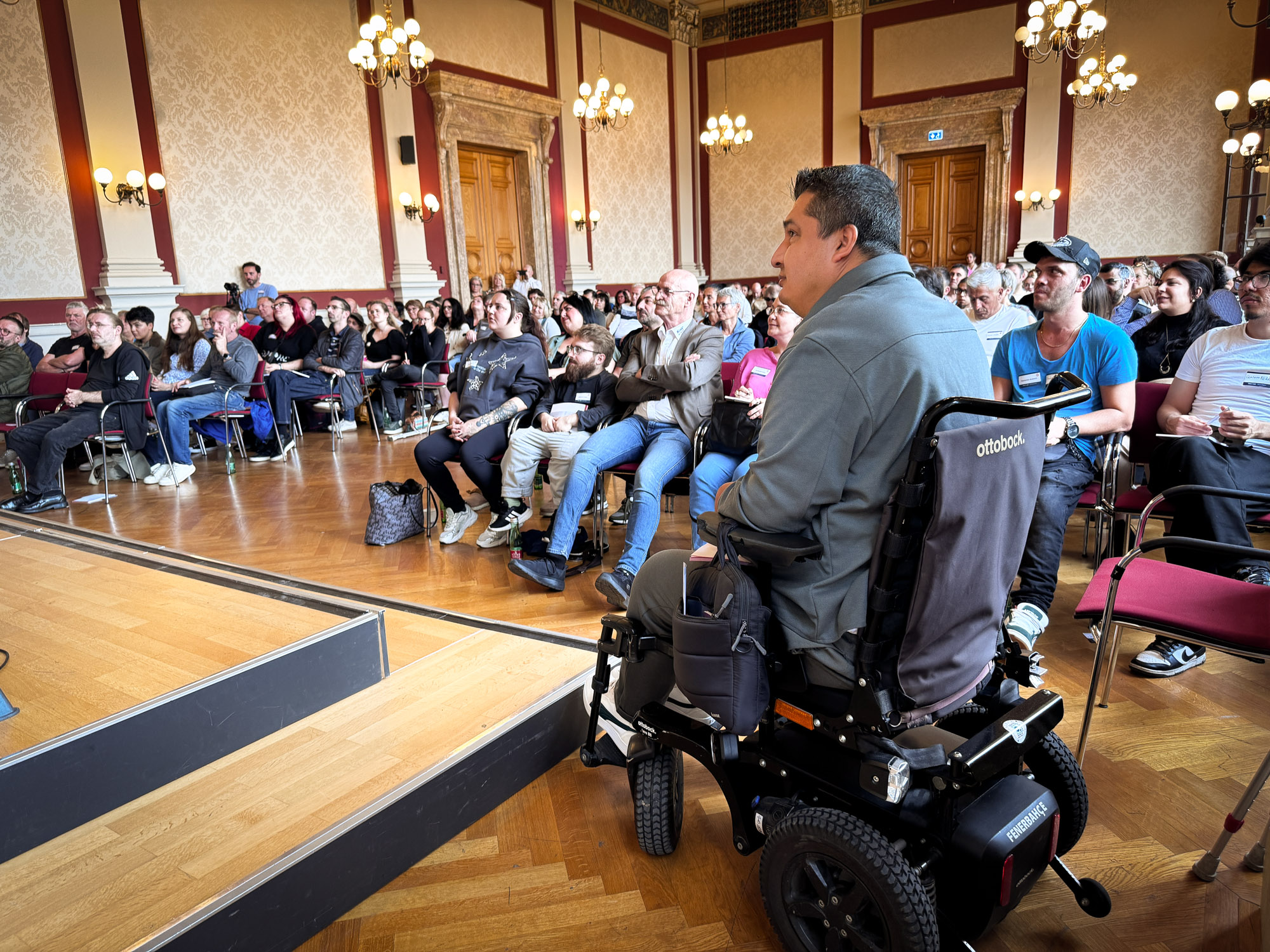 Ein Raum mit Interessentinnen und Interessenten und im Vordergrund ein Interessent im Rollstuhl